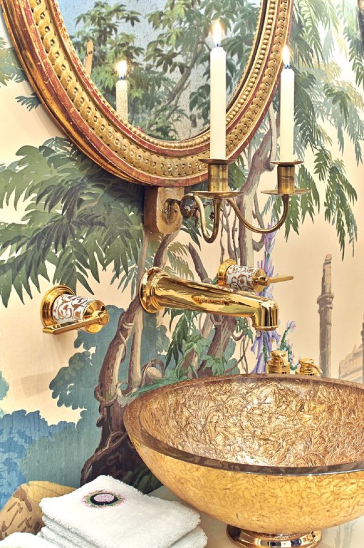 photograph of a powder room with decorative wallpaper, a gold oval mirror, a real candle sconce and a bronze glass bowl sink, visual storytelling by marco joe fazio