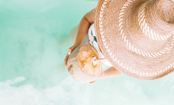An aerial view of a woman with a sunhat standing in the ocean and drinking from a coconut