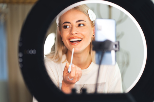 Woman applying lip gloss while recording beauty content with ring light for social media.