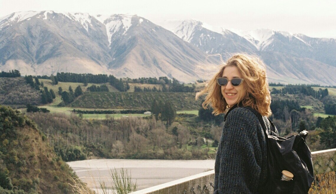 Smiling woman with backpack and sunglasses hiking with scenic snow-capped mountains in the background.