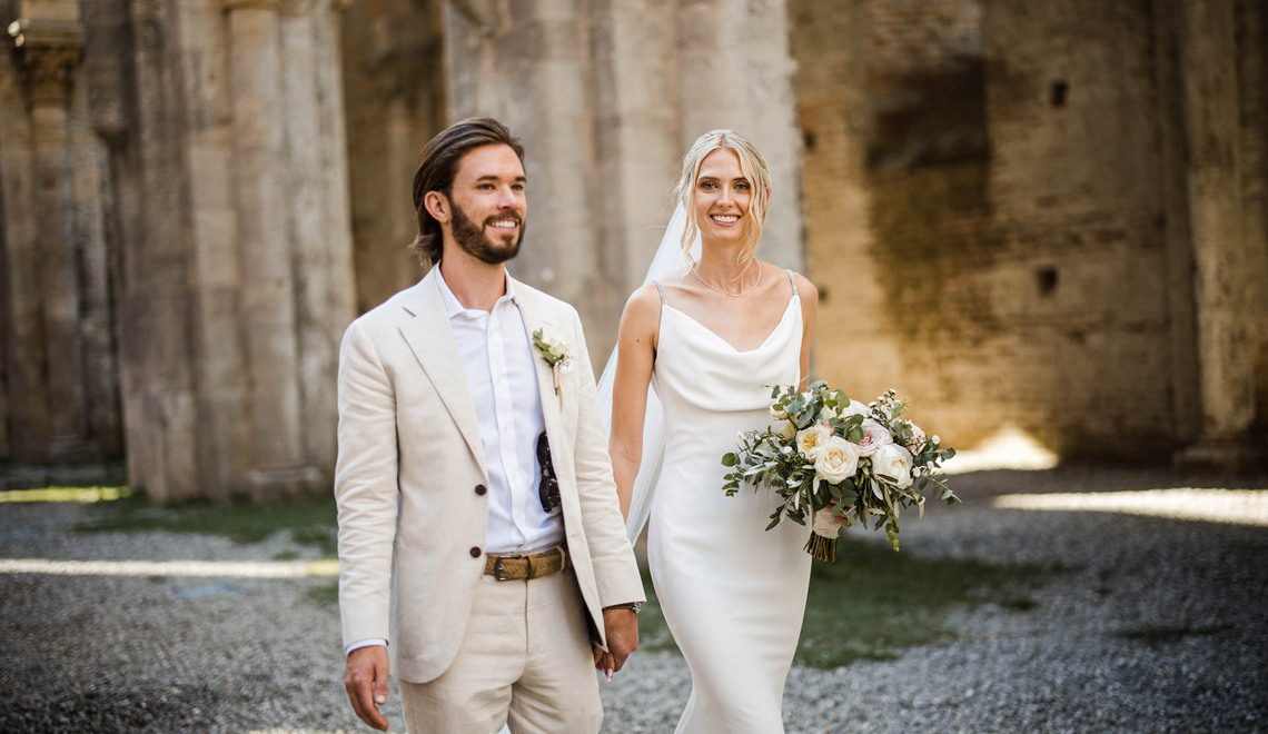 Groom in a beige suit and bride in a white wedding dress holding hands outdoors