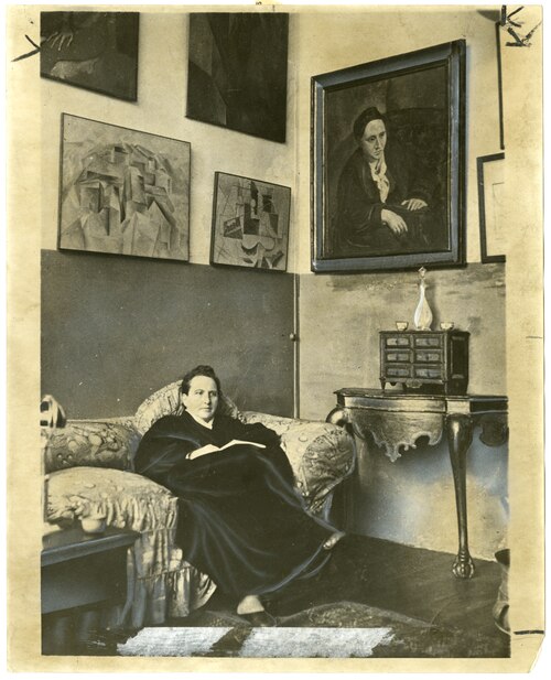 Gertrude Stein sitting on a sofa in her Paris studio, with a portrait of her by Pablo Picasso, and other modern art paintings hanging on the wall behind her, 1930 | Photo via Wikimedia Commons