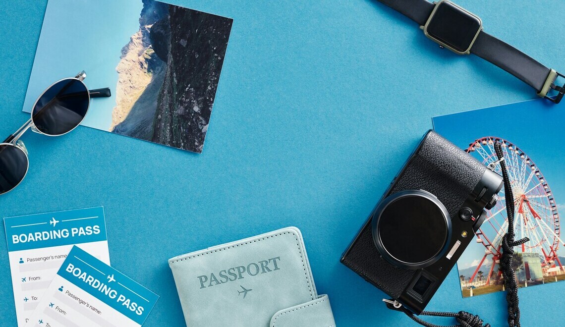 Flat lay of travel essentials including camera, passport, boarding passes, and sunglasses on blue background.