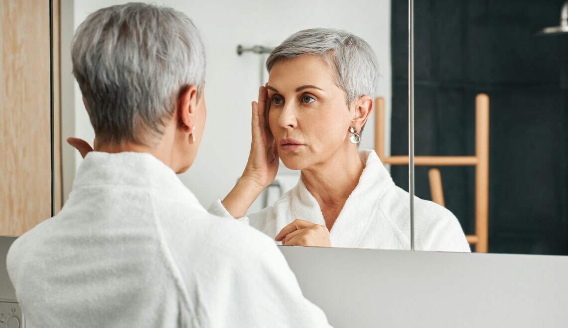Mature woman with short gray hair in a bathrobe looking in a bathroom mirror