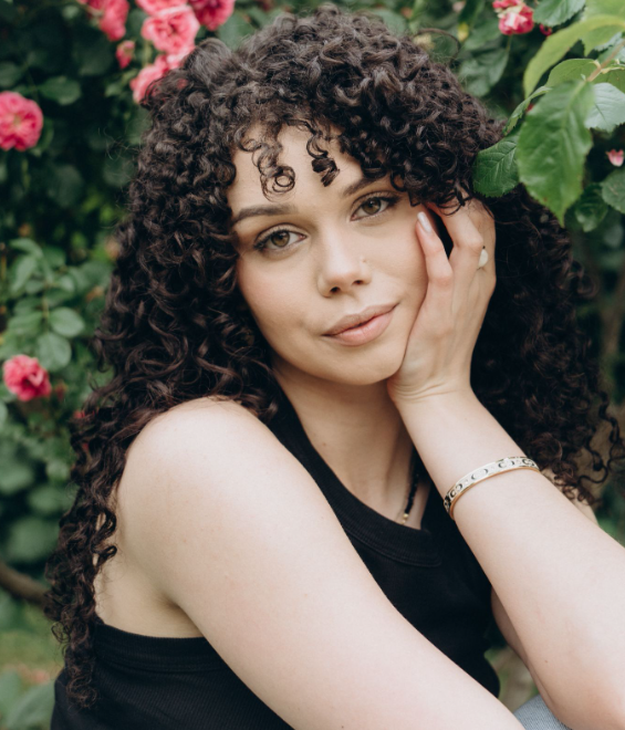 woman with dark curly hair posing outdoors in front of a blooming pink rose bush