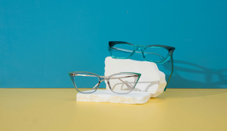 Two pairs of cat-eye eyeglasses displayed on white stone blocks against a blue and yellow background