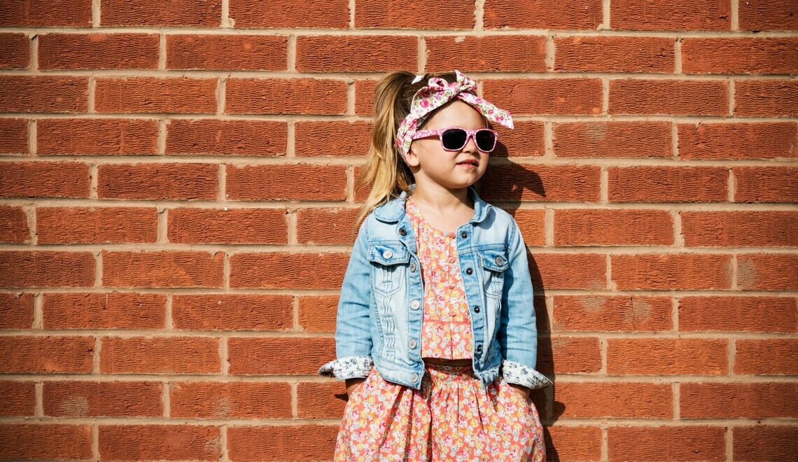 Little girl in a denim jacket, sunglasses, and floral headband leaning against a red brick wall.