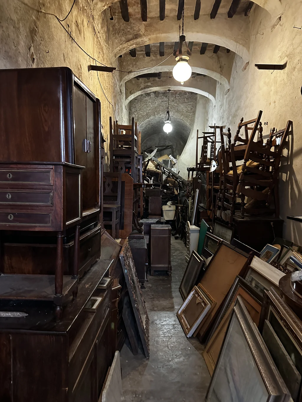 vaulted medieval stone corridor stacked with antique chairs and gilded frames at fiera antiquaria in arezzo